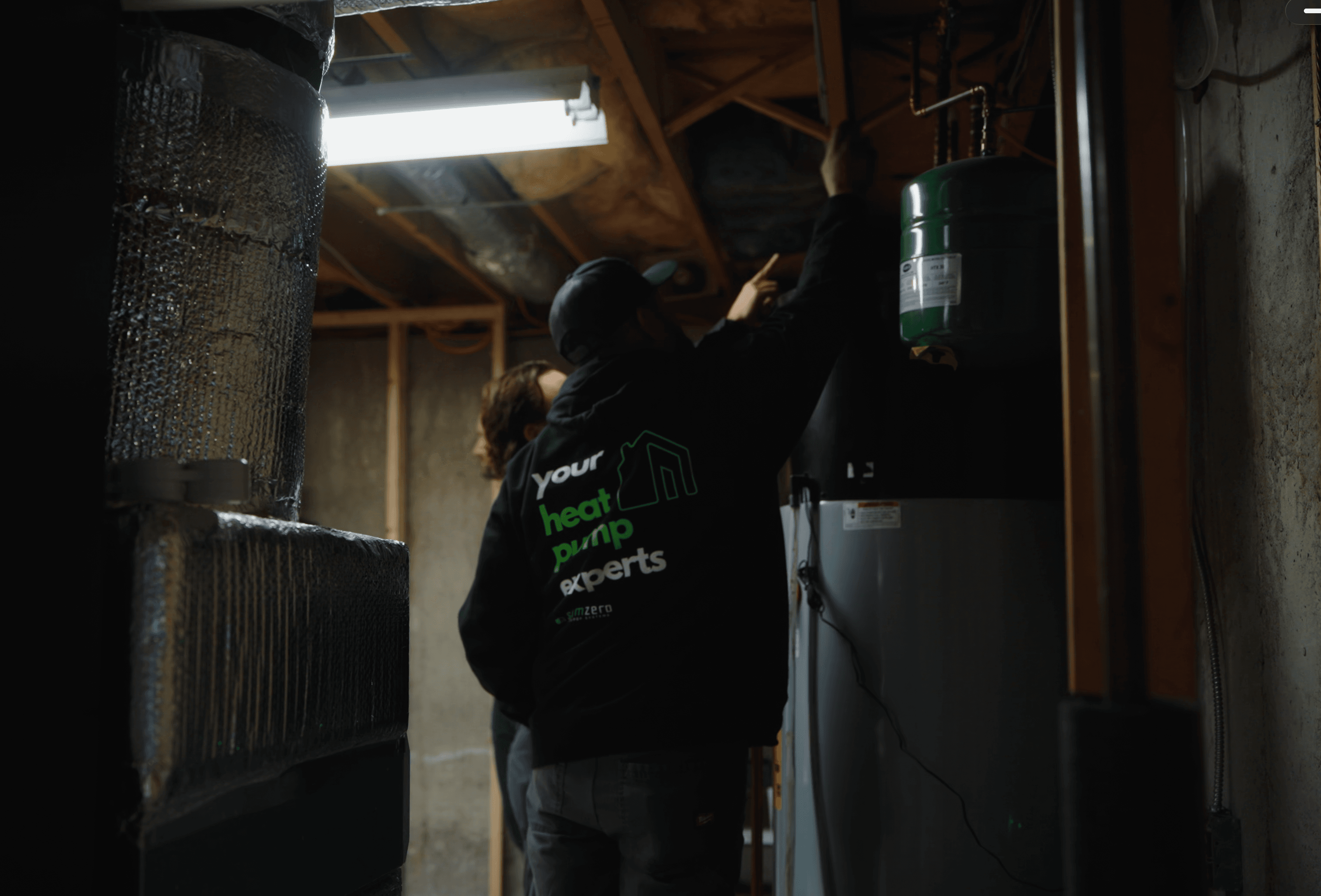 A SumZero technician in a 'your heat pump experts' hoodie walking a homeowner through their basement.